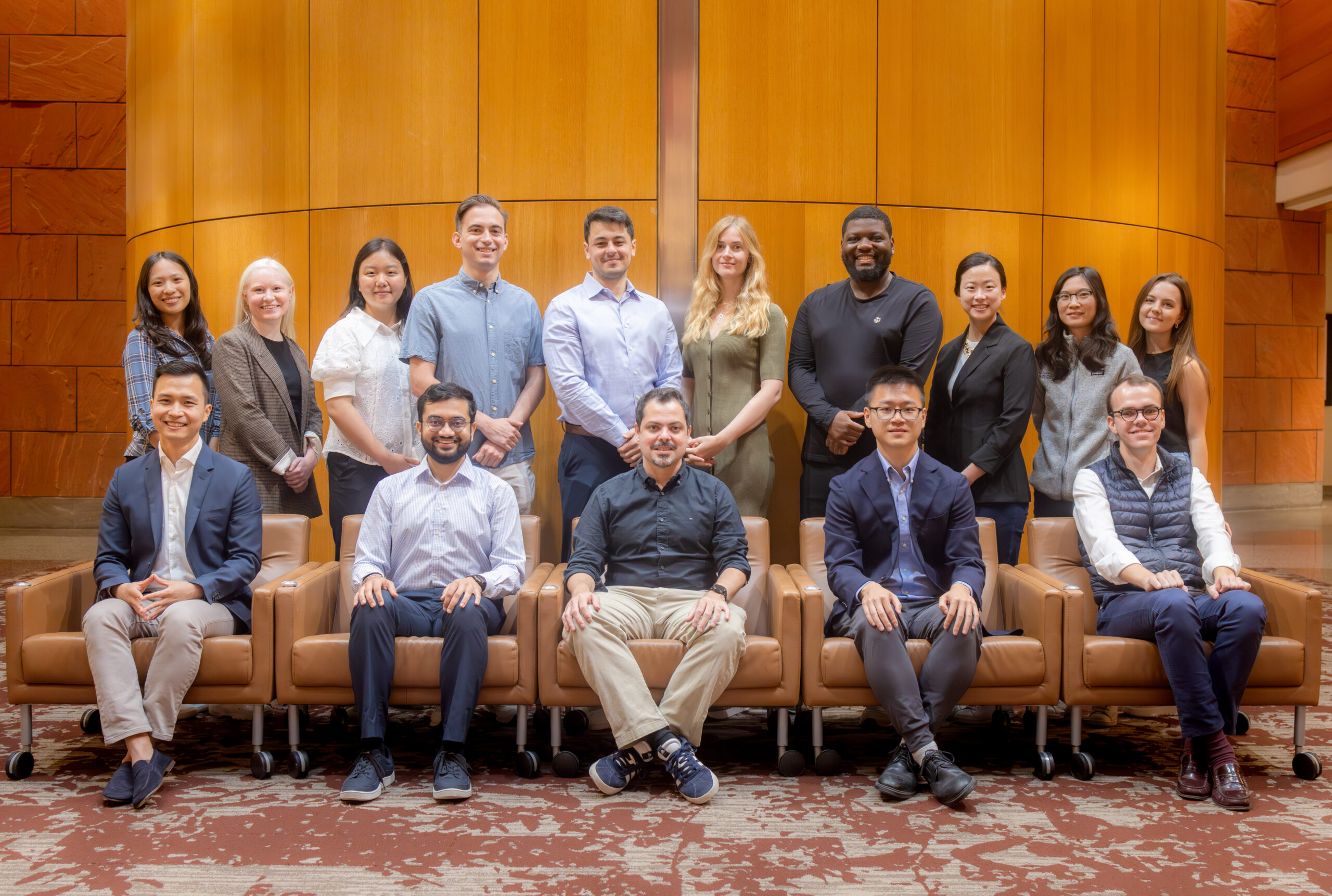 A group of people professionally dressed, posing together indoors in front of a wooden-paneled wall.